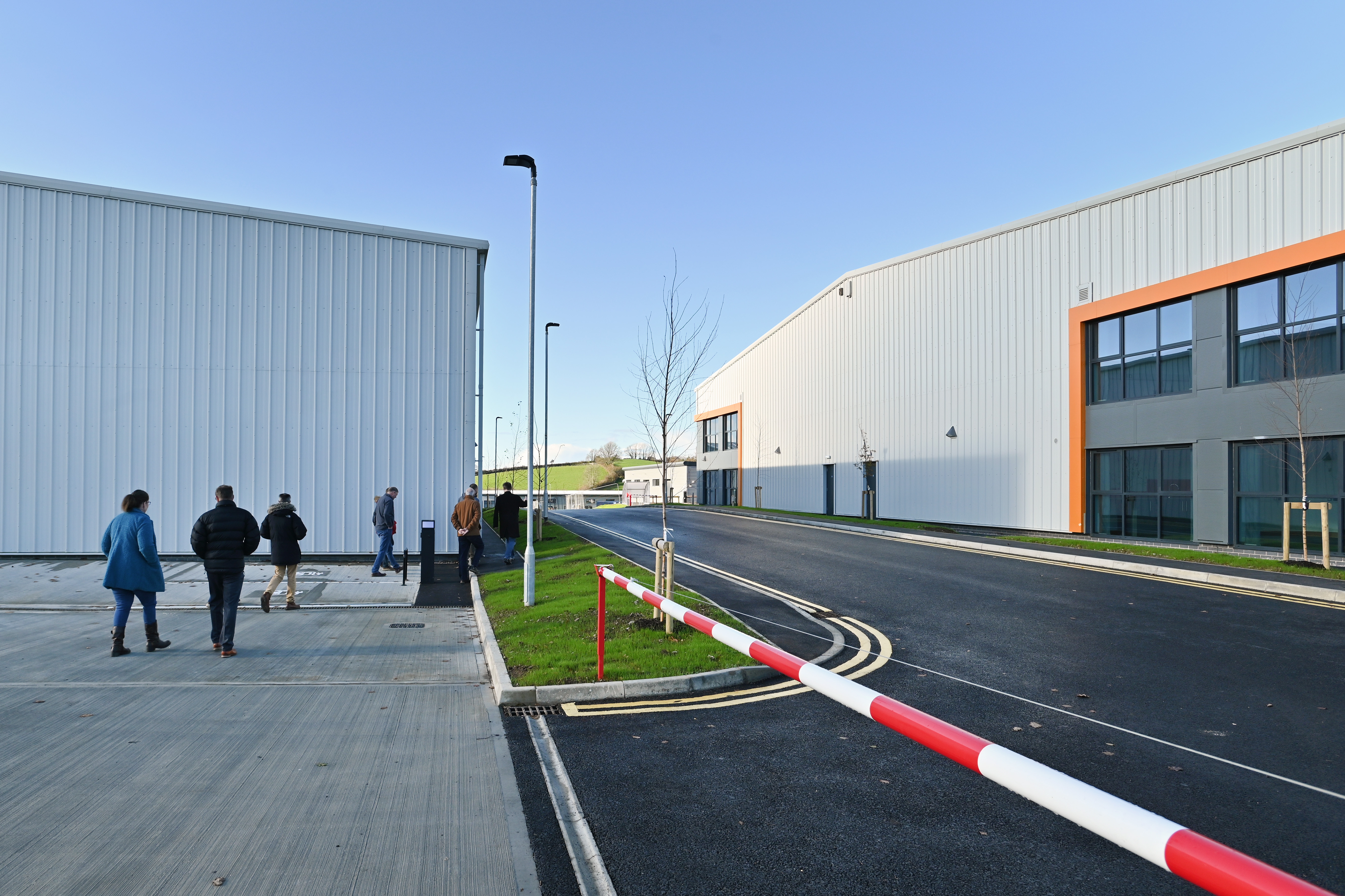 A wide view of a newly developed business park with large modern industrial buildings on both sides of a clean roadway. Several people walk along a paved walkway on the left, while the buildings feature light grey cladding and orange-framed windows. The scene is bright and clear, suggesting a calm day, with streetlights, small planted trees, and a barrier arm visible in the foreground.